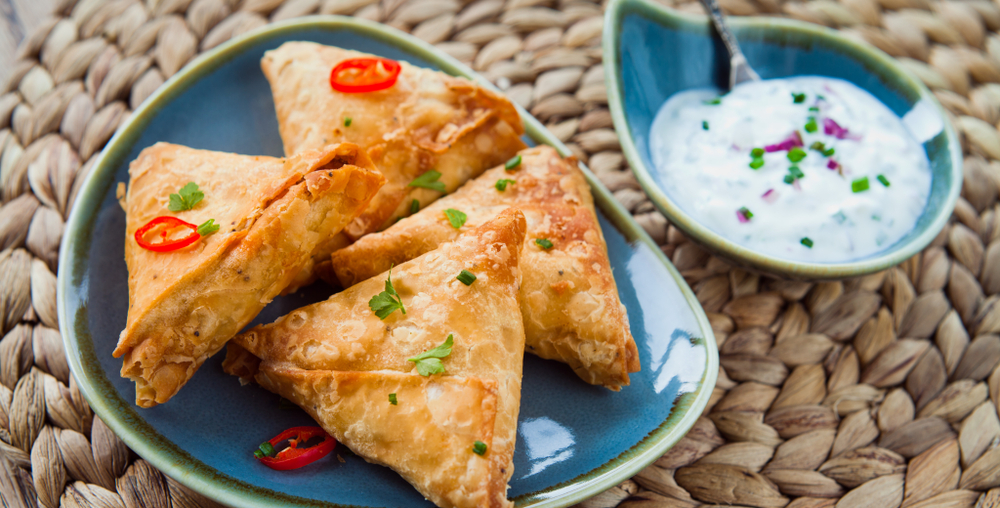 Sambusa on a plate. There are hour on a blue plate with parsley and chilli on top. There is a white dipping sauce nest to it. 