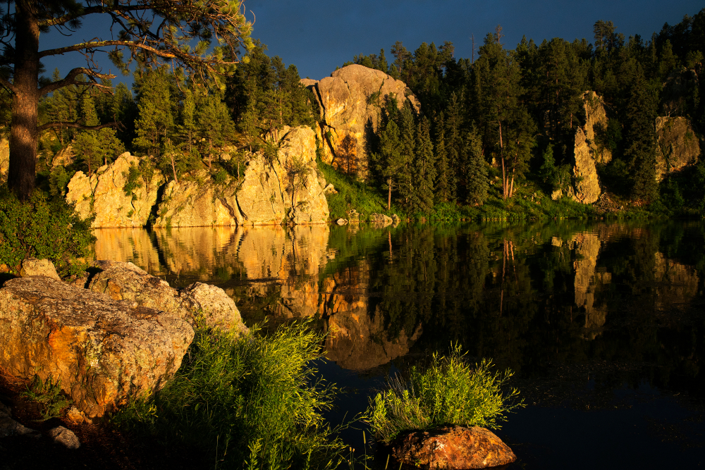 A lake surrounded by huge rock formations and trees as the sun is setting and casting shadows on the rocks. 