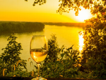 A glass of white wine with a river in the background as the sun is setting, similar to what you'd find at wineries in Missouri.