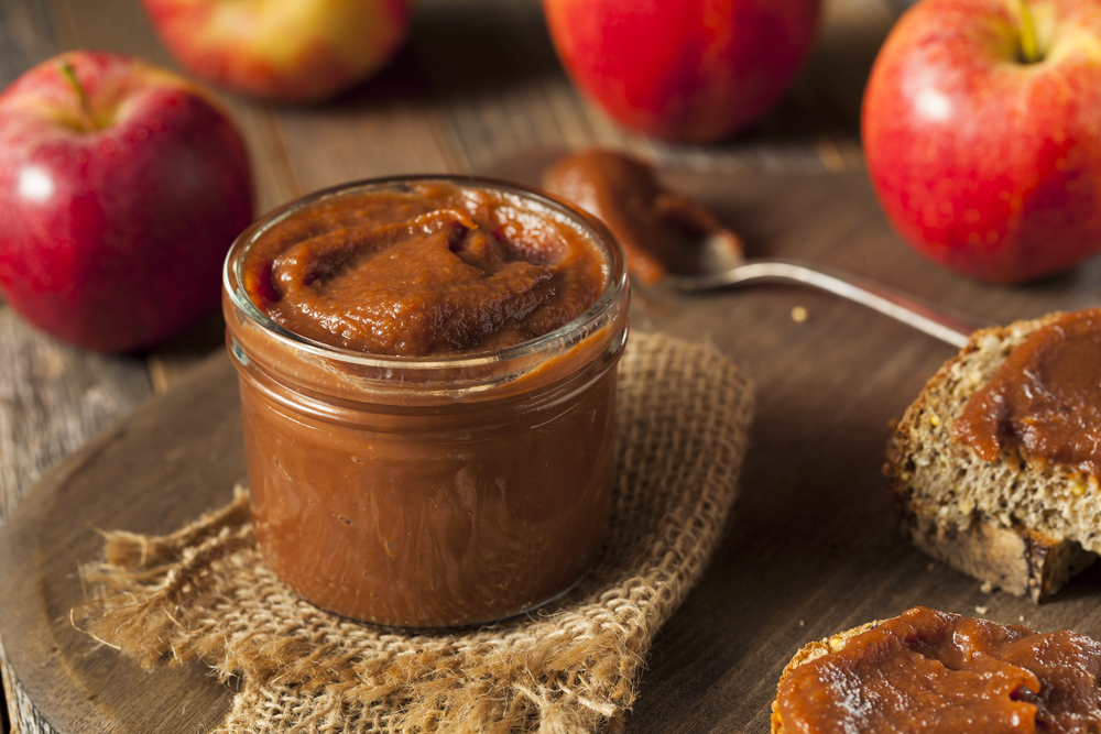 A jar of homemade apple butter with toast that has apple butter on it and some apples on the table. 