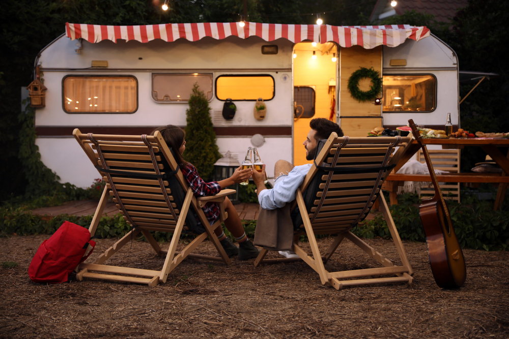 Young couple toasting with bottles of beer near trailer. Camping season. They in front of a camper sat in chairs and a guitar is by the chairs. 