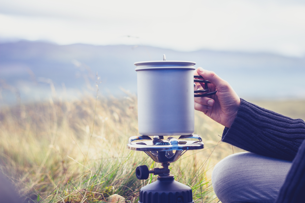 Woman boiling water on portable camping stove. She is wearing a blue cardigan and is in a grass field.  