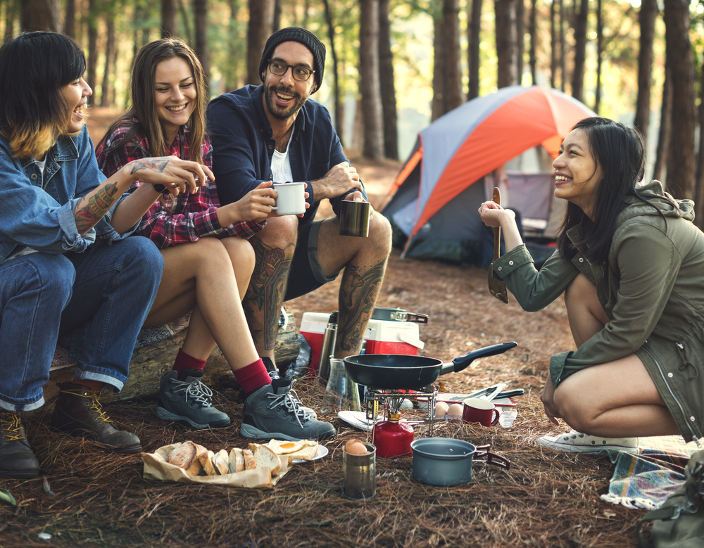 Friends sat around a camp fire drinking and eating and laughing. They are coooking on a stove and their is a tent behind them 