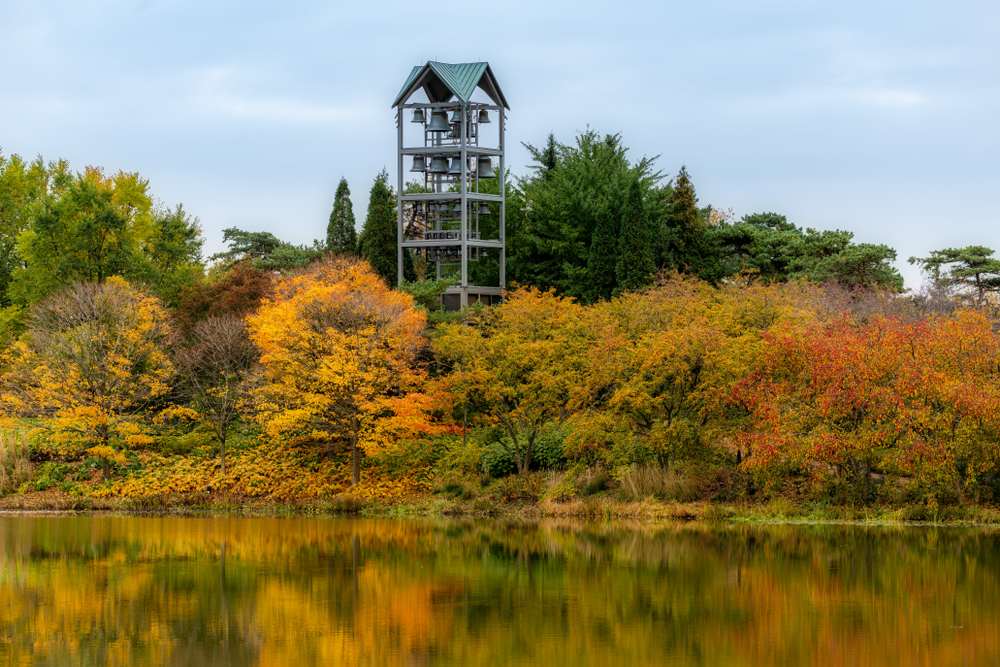 A bell tower on the edge of a lake surrounded by trees at the Chicago Botanic Garden. The trees have orange, red, yellow, and green leaves. 