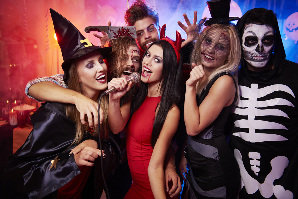 A group of young adults dressed up in costumes, holding a microphone, at a Halloween Costume Party. 