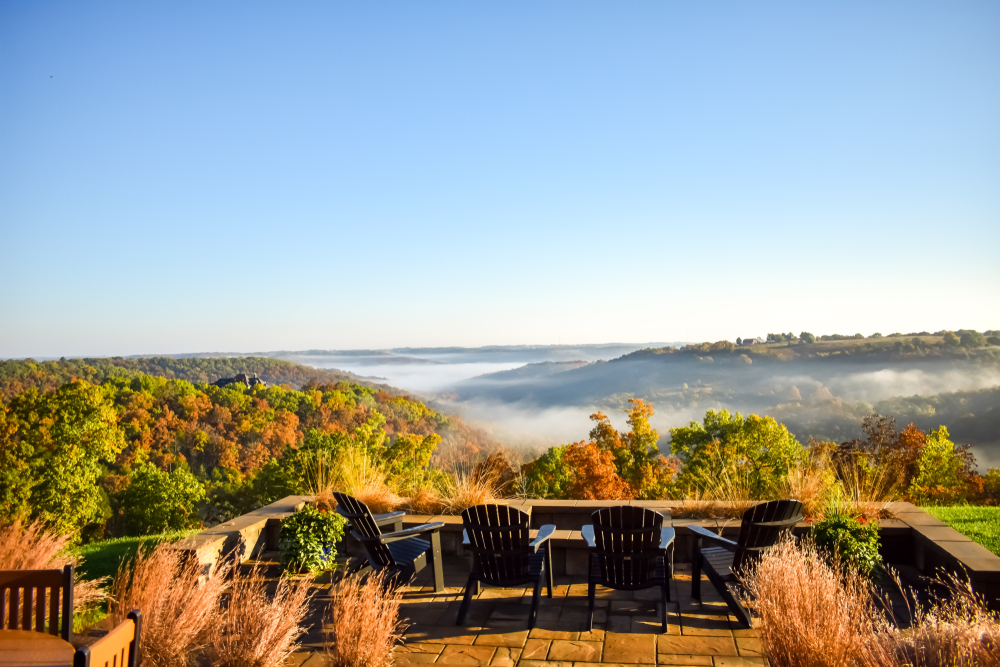 A patio with chairs that are overlooking the Ozarks on a beautiful day in the fall in Missouri. You can see the trees have green and orange leaves. 