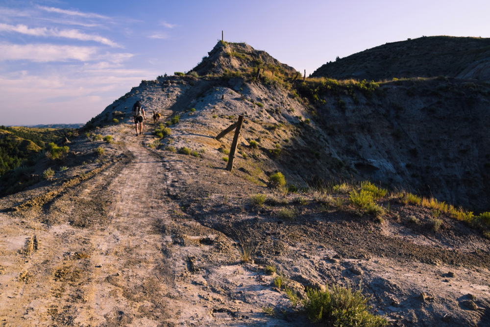 A man and dog hike across Devil’s Pass in late afternoon light. Maah Daah Hey Trail, North Dakota.