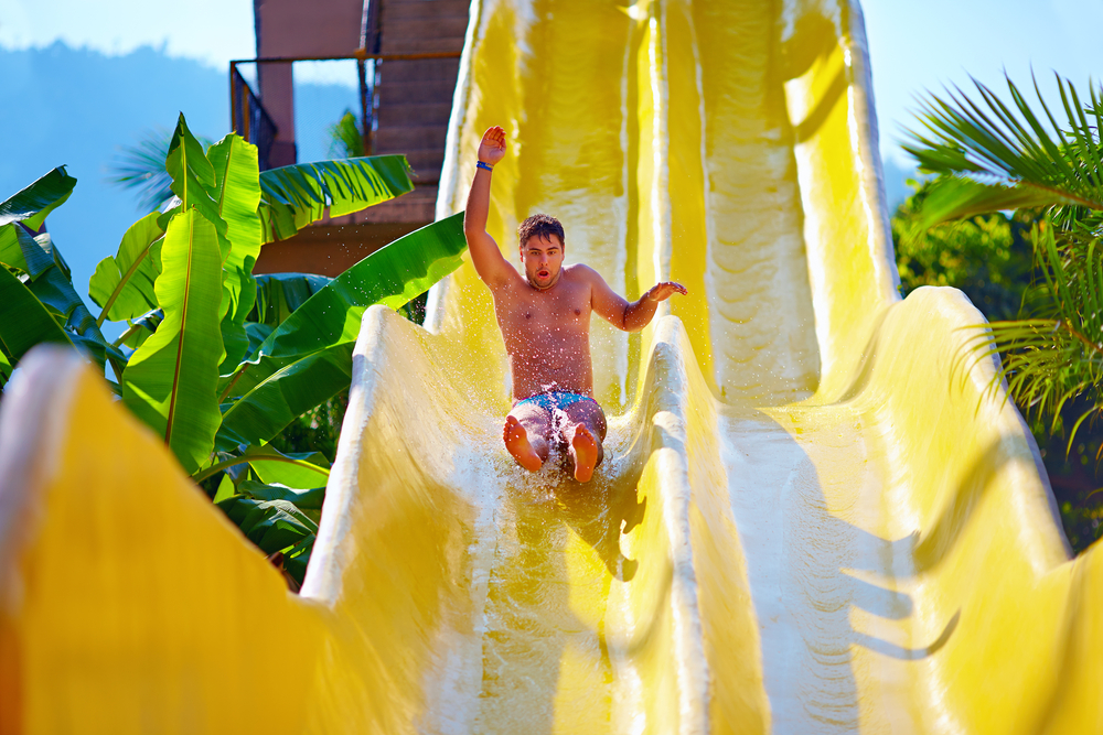 A man going down a steep yellow waterslide outside near some palm trees. Its similar to what you'll find at Wisconsin Dells waterparks. 
