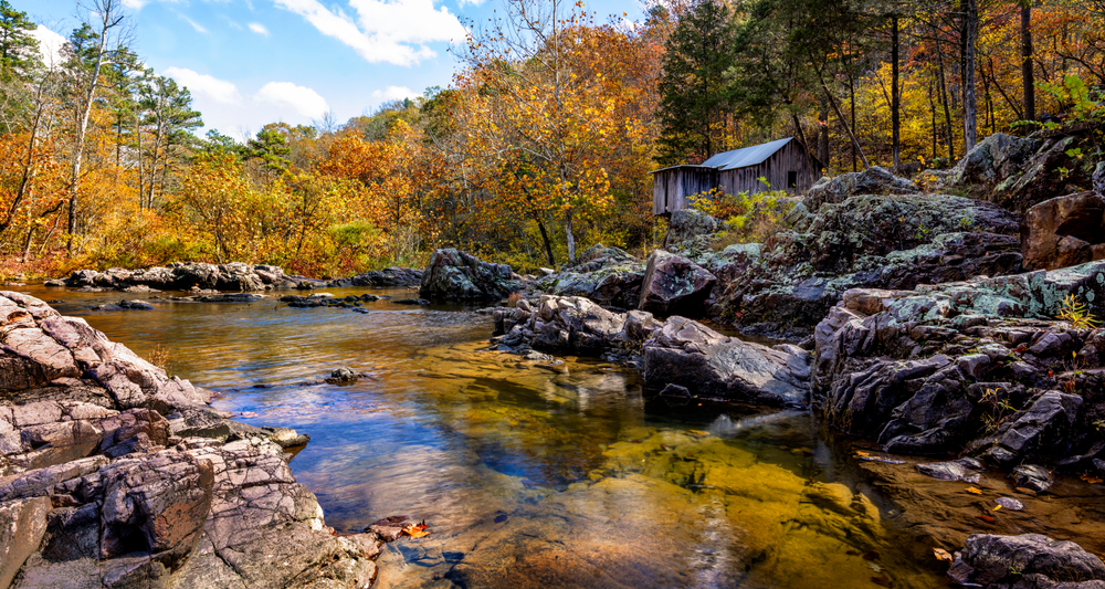 A river with a rocky shore in the Mark Twain National Forest with a small spring cabin on the shore. You can see yellow and orange fall foliage sorrounding the river. 