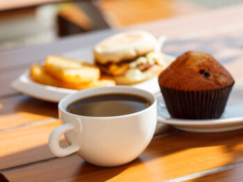 Coffee in white cup with muffin and breakfast muffin sandwiches in background. Restaurants in Evansville