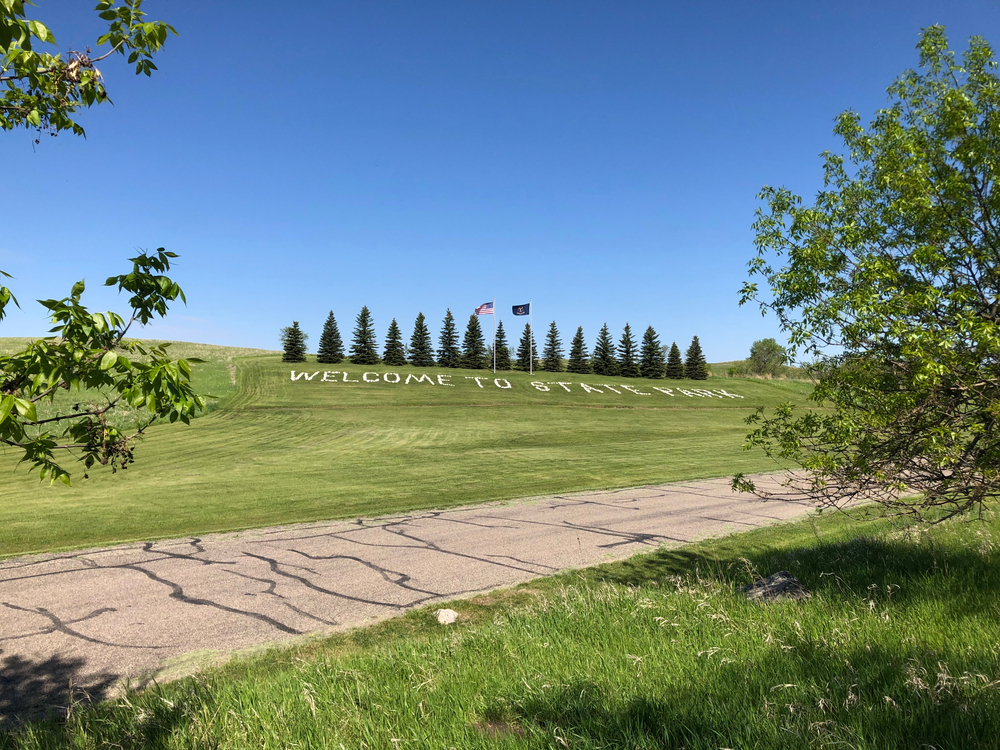 The entrance to a state park in North Dakota with green open spaces and trees. Welcome to State Park is written on the ground. 