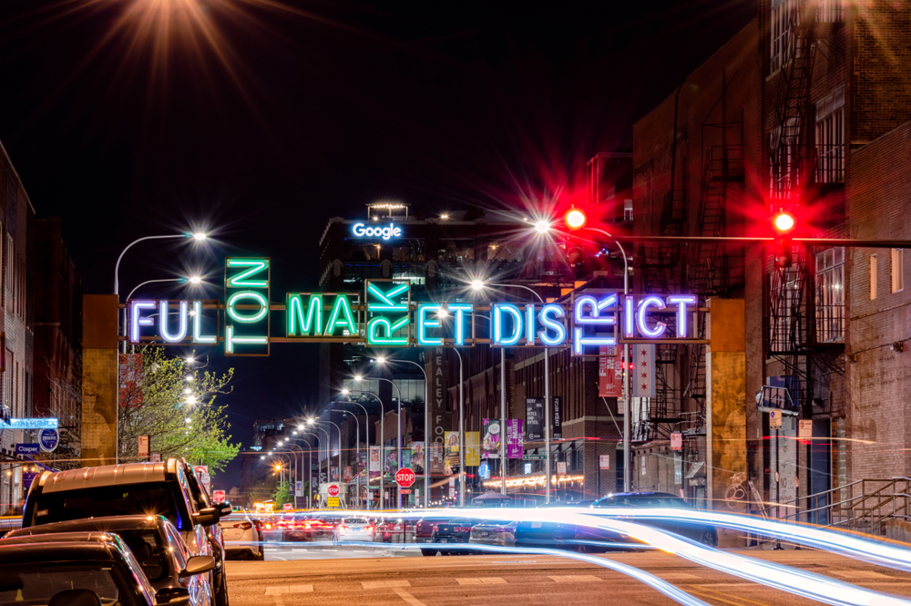 Night shot of the Fulton Market District sign lit up in different colors over a street with cars.