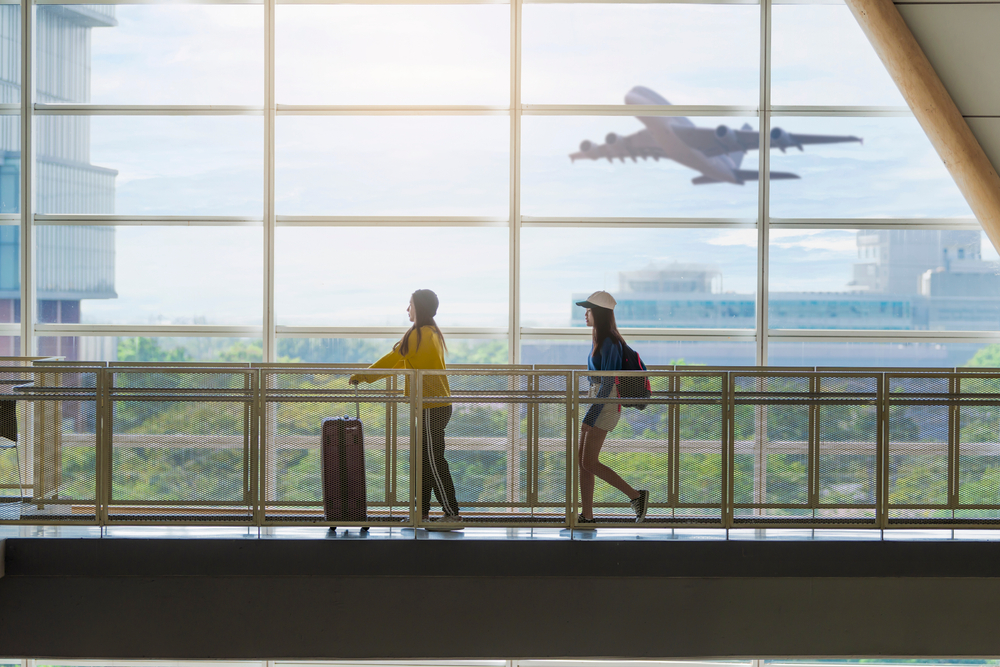 Two people walking in front of a huge window at an airport and you can see a plane taking off outside. 