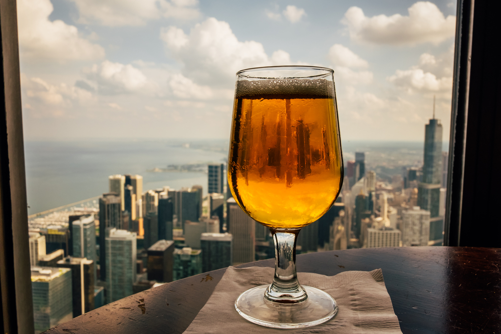 A glass of beer on a table in front of a window overlooking the Chicago skyline on a nice day. 