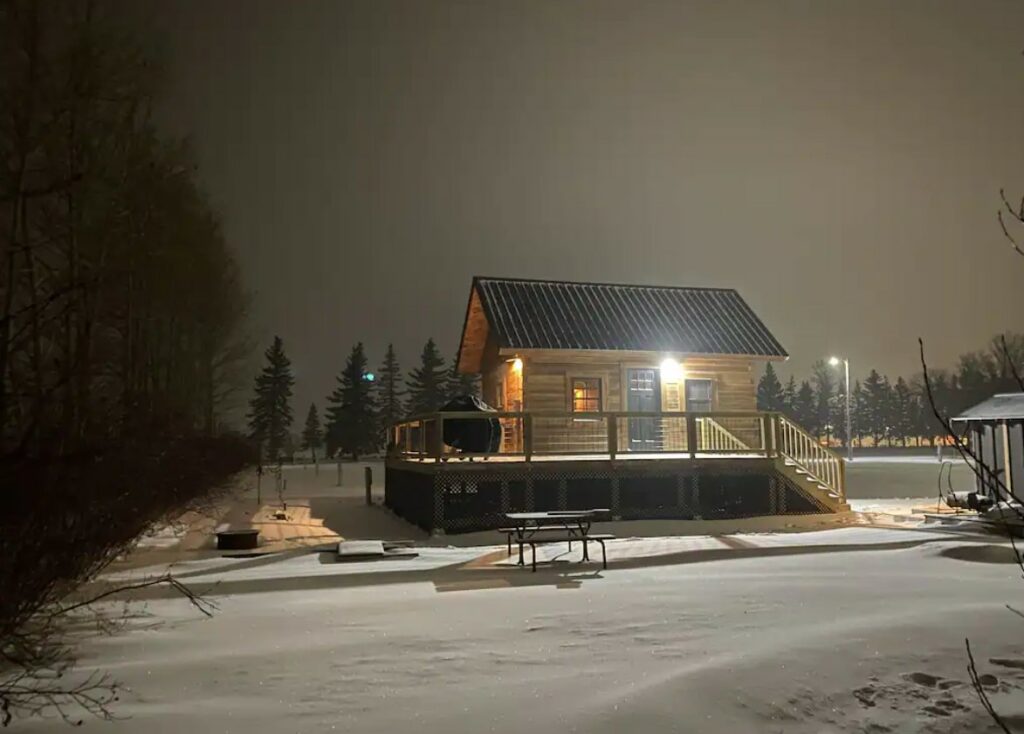 The exterior of a classic log cabin with a wrap around porch on a winter night. The cabin is surrounded by a yard that is covered in snow and there is a light on the porch and a light on in the cabin. 