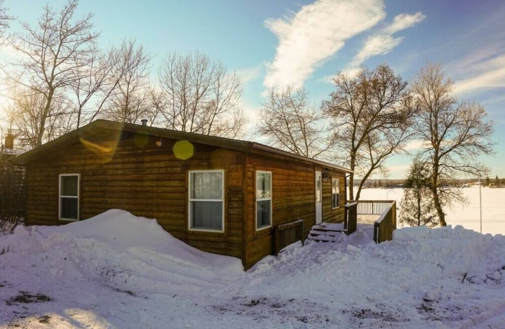 The exterior of a log cabin from the side that is surrounded by snow and on the shore of a lake, one of the best cabins in North Dakot