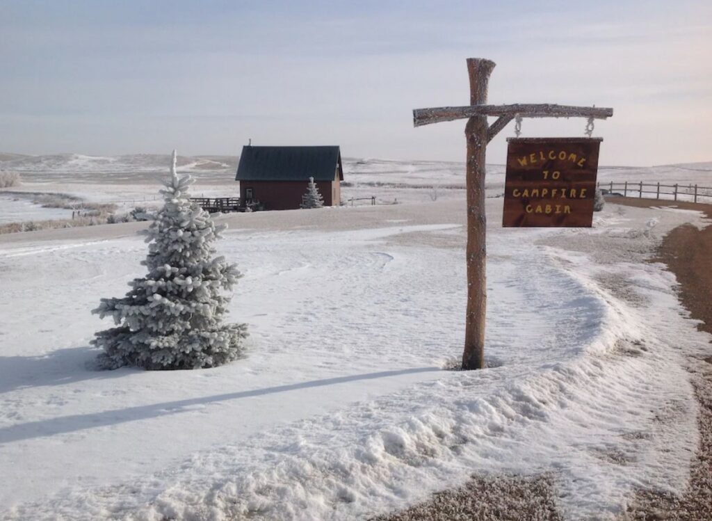 One of the best cabins in North Dakota on the prairie that is coveer4ed in snow. There is a sign welcoming you to the cabin and you can see it in the distance