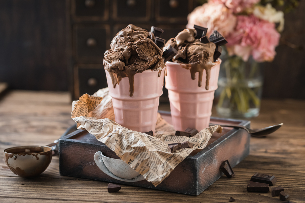 Chocolate ice cream sundae in two pink cups on a block of wood. There si chocolate and an ice cream scoop on the table. Pink flowers are in the background. 