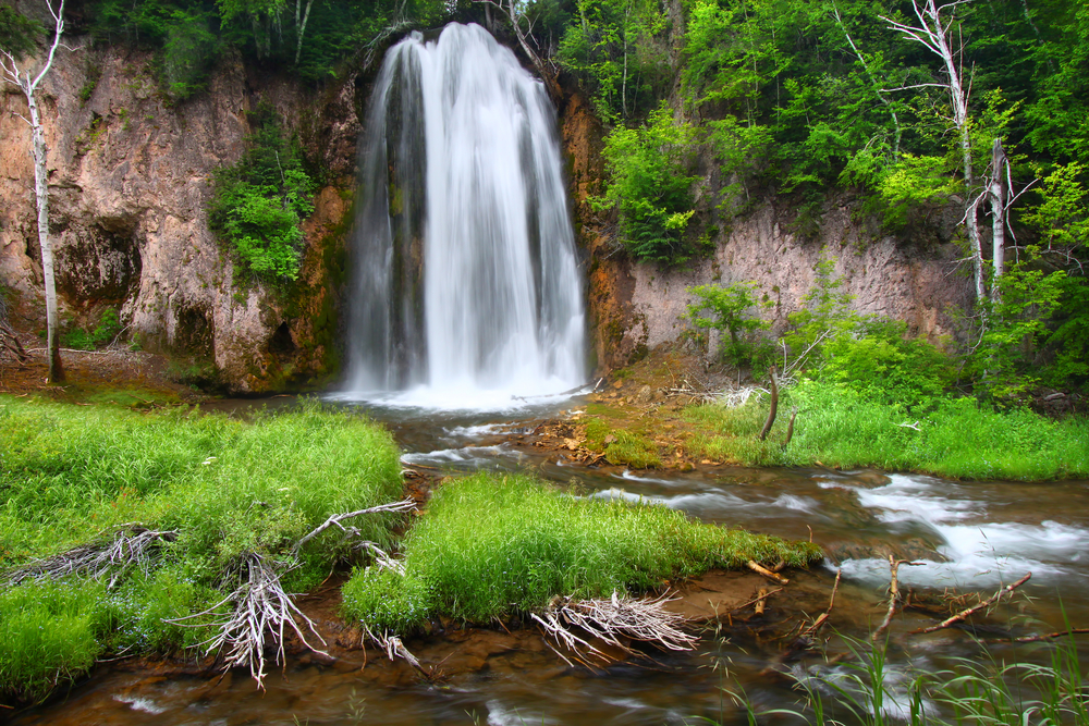 The spearfish falls waterfall on a summer day surrounded by green foliage, rock formations, and a creek. 