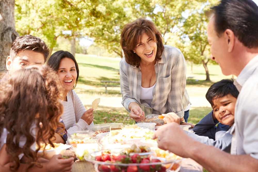 Happy multi generation family having a picnic in a park. There is food on the table and the mum is stood up