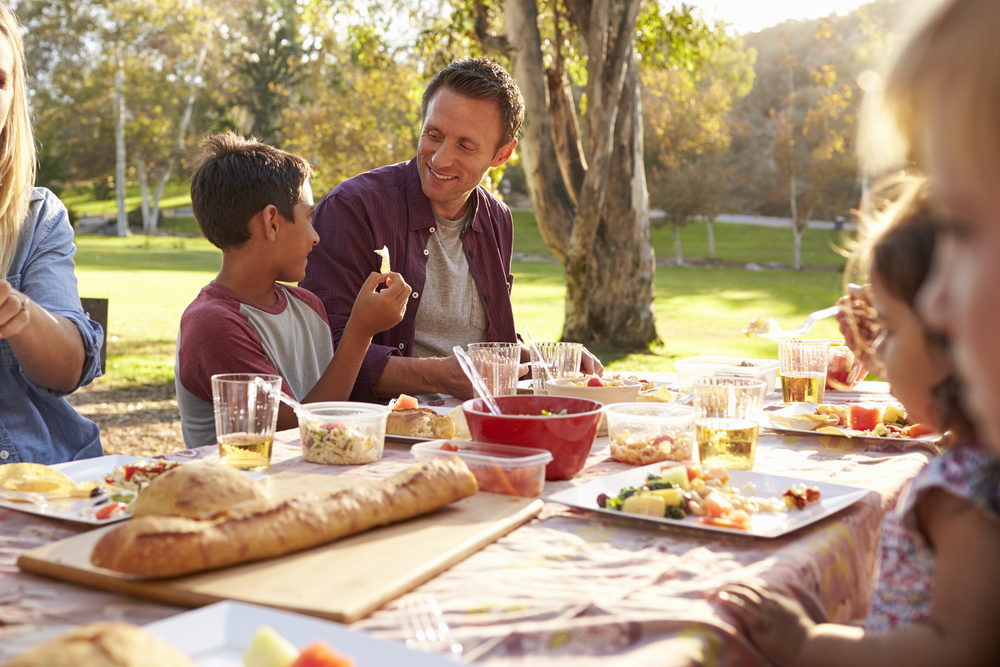Two families having a picnic at a table in a park, close up. The table is full of food 