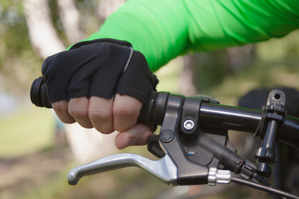 close up of a gloved hand holding the handlebars of a bike. The glove is black and the shirt green. 