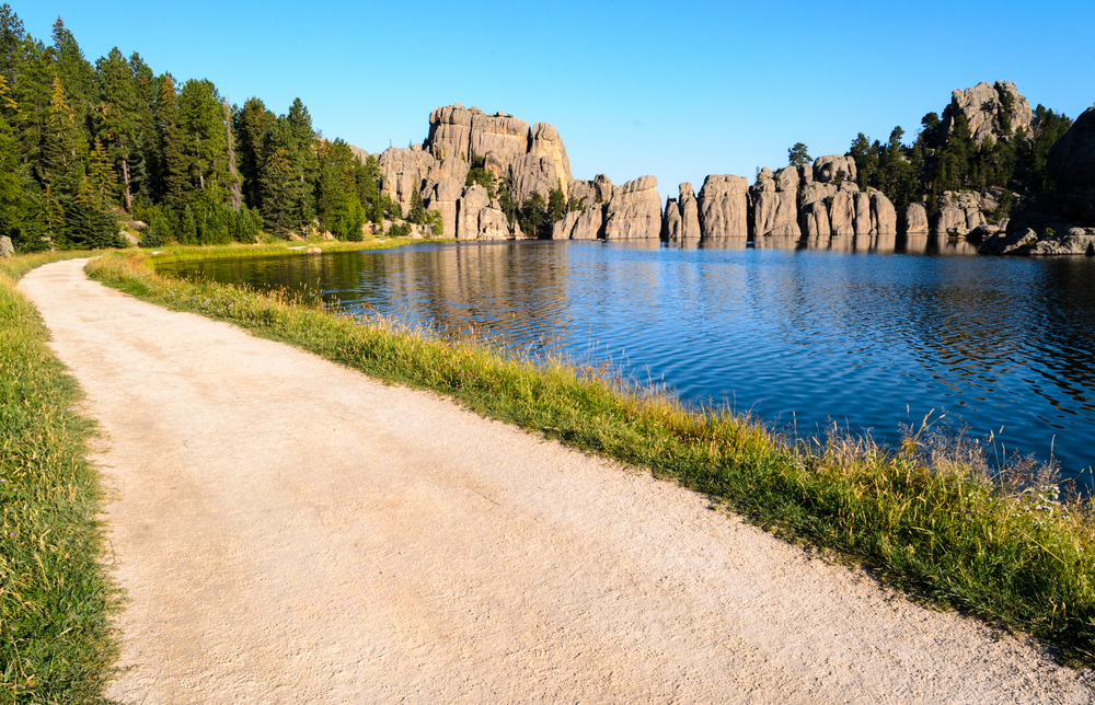 Hiking is one of the things to do in the Black Hills. The picture shows a trail beside a lake with rocks in the background.   