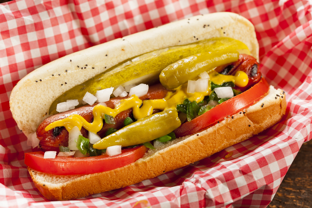 Chicago Style Hot Dog with Mustard, Pickle, Tomato, Relish and Onion it is a container with a red gingham napkin