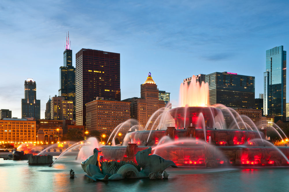 Buckingham Fountain in Grant Park. It is all lit up at night and the city skyline is lit up in the abckground.