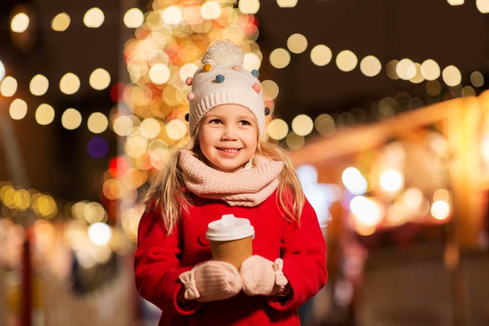 A young girl holds a cup of hot cocoa with a light display in the background.