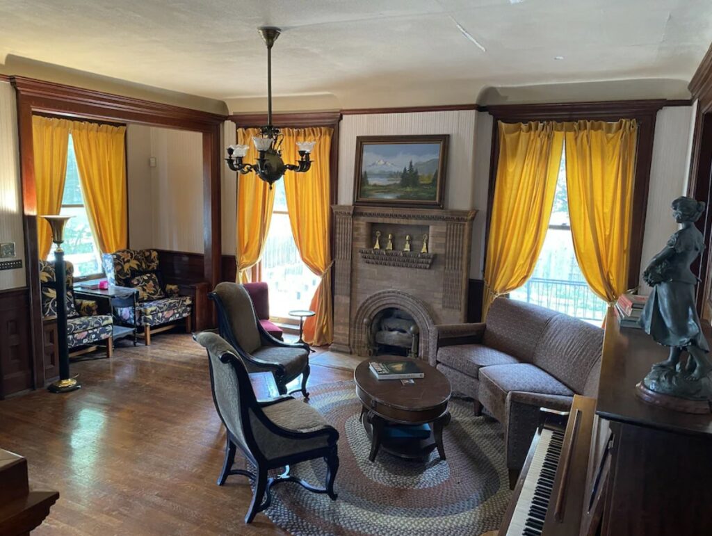 The living room of a historic airbnbs in Kansas City Missouri with a large brick fireplace, large windows with yellow curtains, and a piano. 