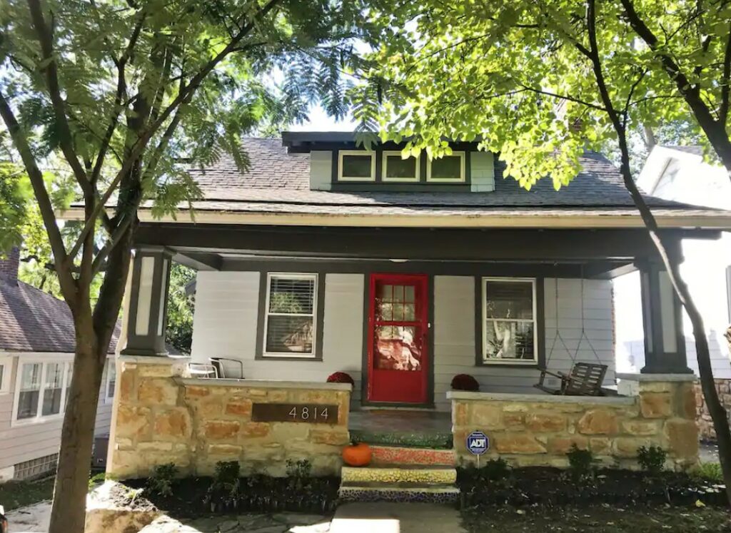 The front exterior of a small bungalow house with a stone front porch and a bright red door
