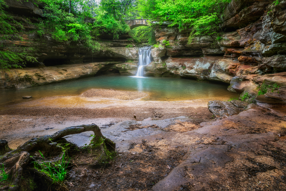 A pond of water that is fed by a waterfall surrounded by rocks and lush greenery, one of the best caves in Ohio