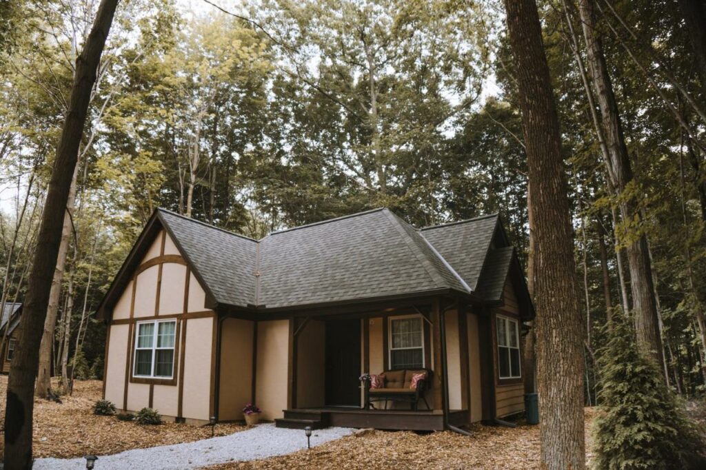 A small light brown chalet in the woods that has a small front porch