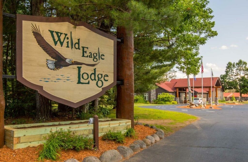 The front entrance to a lodge that has a sign with an eagle painted on it that says 'Wild Eagle Lodge' surrounded by pine trees