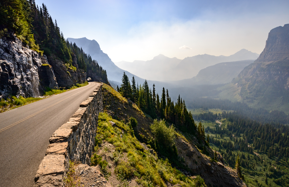 Portion of road going along a mountain edge with more mountains and forests in the distance.