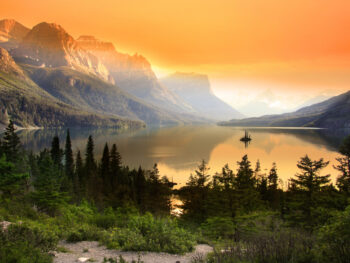Orange sunset sky in background, glassy lake, and evergreen trees in middle.
