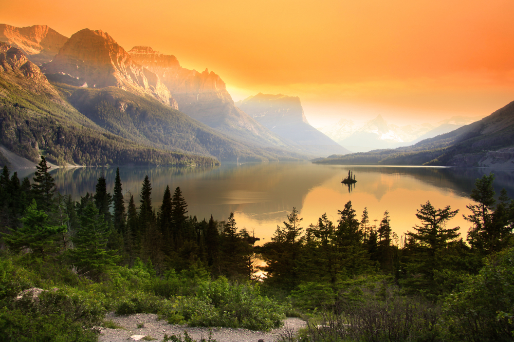 Orange sunset sky in background, glassy lake, and evergreen trees in middle.