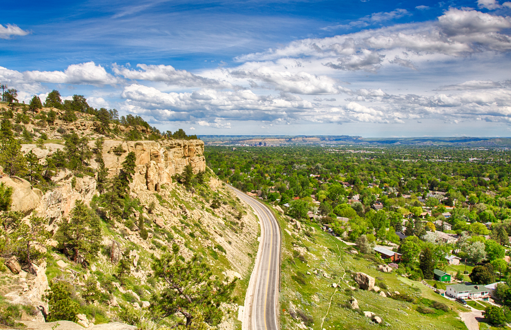 Road winding beside huge sandstone cliffs on one side and a green City on the other side things to do in billings