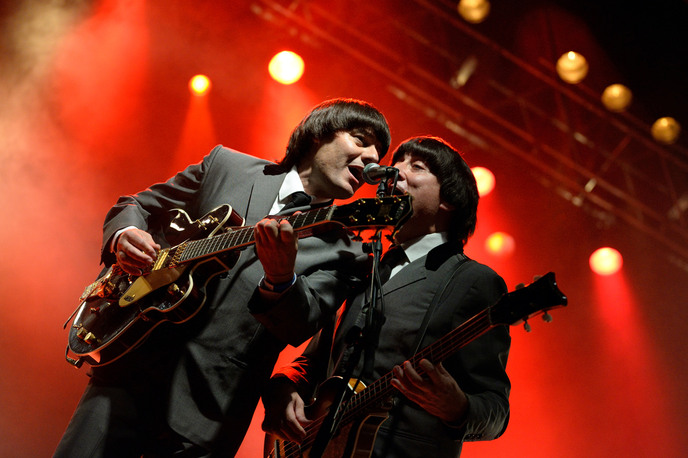 Two men dressed like The Beatles playing guitar and singing on a stage with red lights