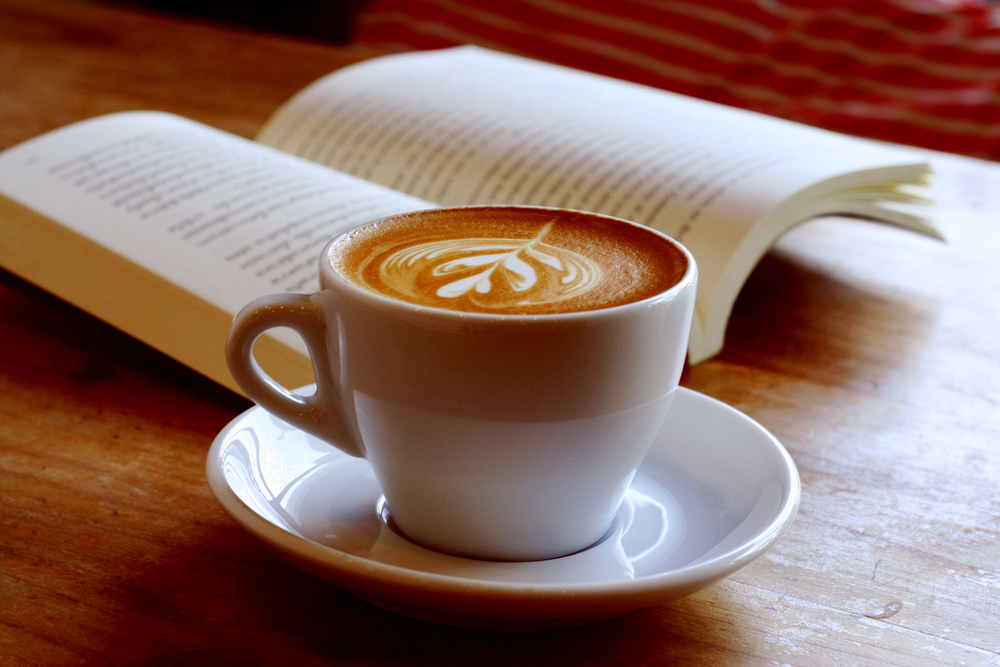 A latte in a white and tan mug on a table next to a book that is open