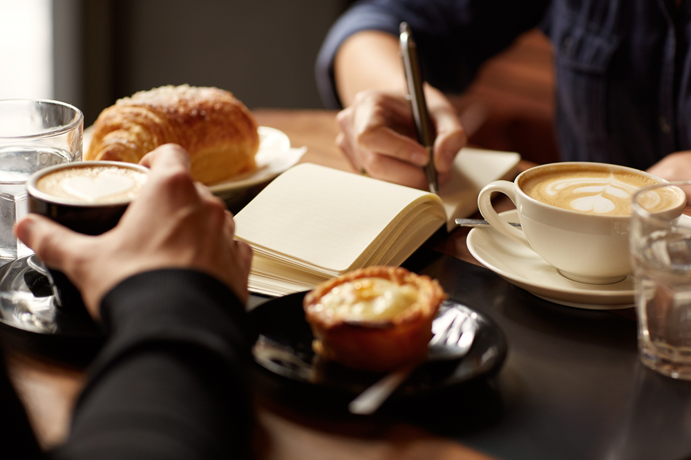 A table with coffee, pastries, and you can see the hands of two people, one of them is writing in a notepad