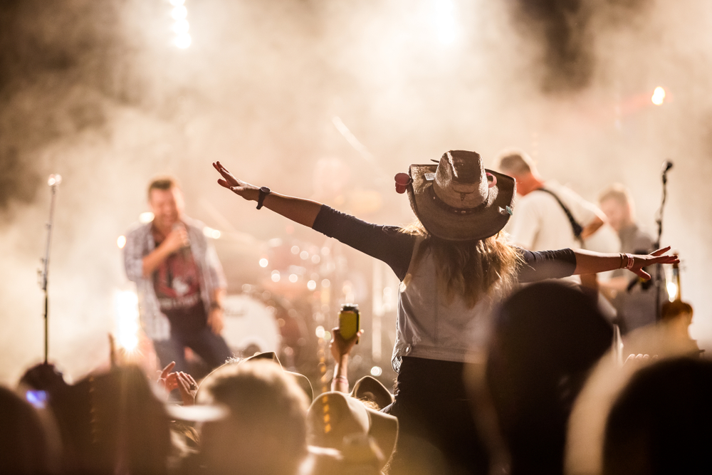 A person on someone's shoulders wearing a cowboy hat with their arms outstretched looking towards a stage where there is a band playing at one of the best festivals in Ohio