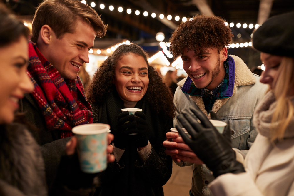 A group of young people standing together under string lights at an outdoor festival holding cups with warm beverages. 