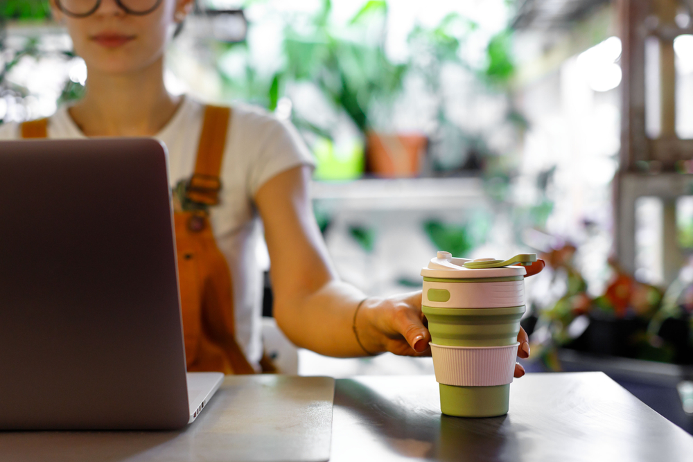 A woman in a coffee shop full of plants working at a laptop drinking out of a reusable coffee cup