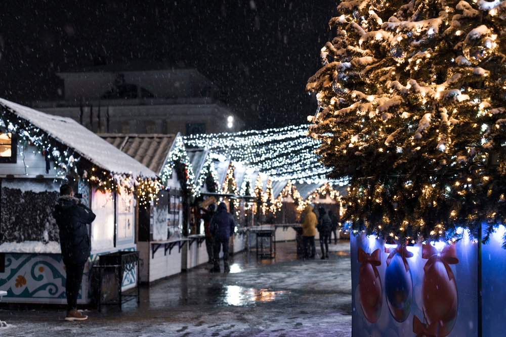 A winter fair at night that has small vendors, string lights, and a large lit up Christmas tree