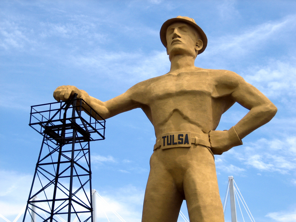A large statue that is a gold color that resembles an oil driller. He has his arm resting on a piece of drilling equipment. 