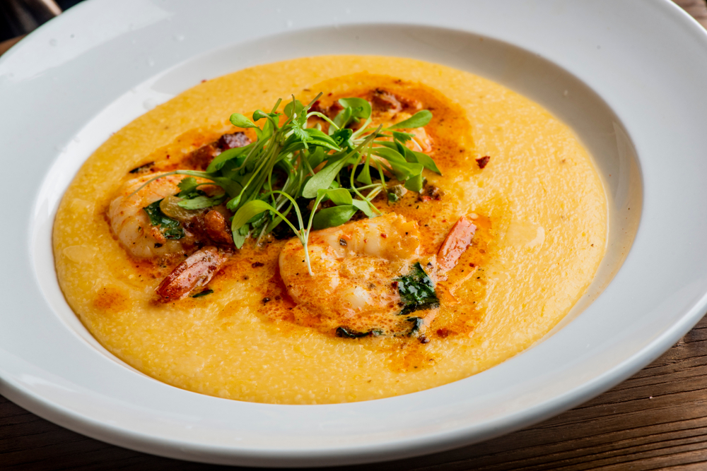 A large white bowl with yellow grits, shrimp, a red sauce, and fresh greens