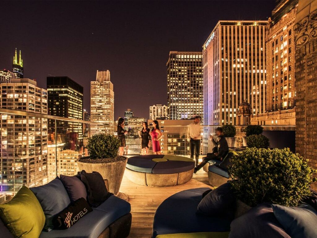 People standing on the rooftop terrace at Virgin Hotels Chicago at night with city views.