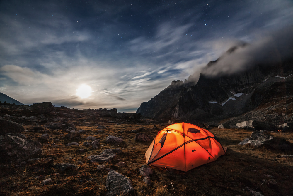 An orange tent glowing from the inside on the side of a mountain at night with the moon shining